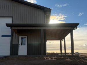A gray metal building with a covered side entrance and white door, photographed in late afternoon sunlight with dirt ground in the foreground.