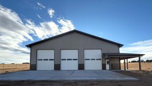 A large metal building with three white garage doors and an attached covered side area, set on a concrete lot under a partly cloudy sky.