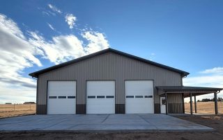 A large metal building with three white garage doors and an attached covered side area, set on a concrete lot under a partly cloudy sky.
