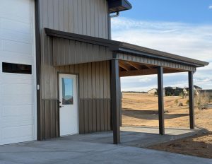 A metal building with a white door, a covered porch area supported by posts, and a concrete walkway; dry grass and houses are visible in the background.