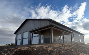 A large gray metal building with three garage doors and a covered entryway sits on a dirt lot under a partly cloudy sky.