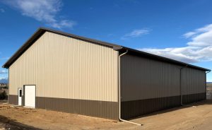A large metal-sided building with a brown and beige exterior stands on a dirt lot under a blue sky with some clouds.