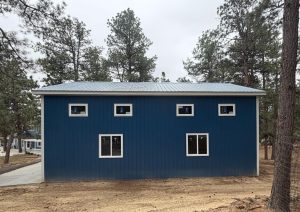 A blue metal-sided building with a row of small square windows above two larger rectangular windows, surrounded by pine trees on a dirt lot.