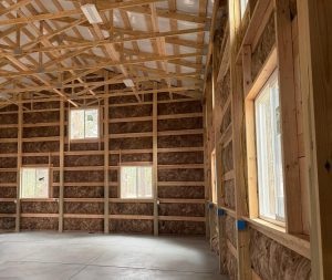 Interior of a building under construction showing exposed wooden framing, insulation in the walls, three windows, and a concrete floor.