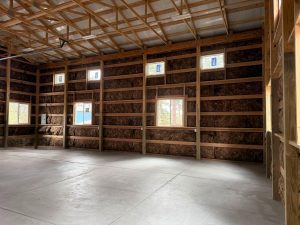 Interior of an unfinished building with exposed wood framing, four small windows, insulation in the walls, and a concrete floor.