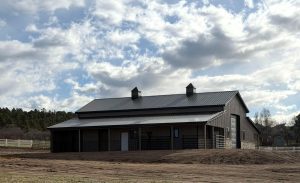 A modern barn with a metal roof and two cupolas sits on a dirt lot under a partly cloudy sky, surrounded by trees and a white fence.