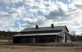 A modern barn with a metal roof and two cupolas sits on a dirt lot under a partly cloudy sky, surrounded by trees and a white fence.