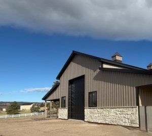 A metal and stone barn with a large black door stands under a partly cloudy sky, surrounded by a dirt lot and a white fence in a rural setting.