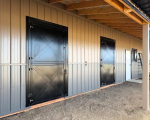 Exterior view of a metal building with two large black barn doors, a white standard door, and a wood-framed roof overhang.