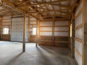 Interior of a partially finished wooden structure with exposed beams, insulated walls, a concrete floor, and a large garage door letting in natural light.