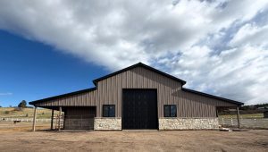 A large, brown barn with a stone lower exterior and central black door stands under a partly cloudy sky in a rural area.