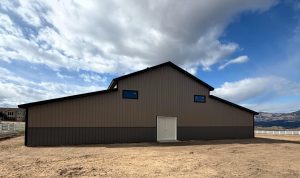 A large, modern brown barn with small windows and double doors stands on dirt ground under a partly cloudy sky, with mountains and a white fence in the background.