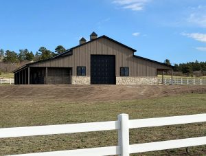 A large barn with a stone and metal exterior, black double doors, and cupolas on the roof, surrounded by a white fence and open grassy area.