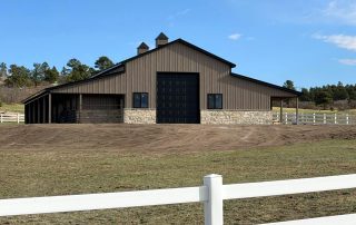 A large barn with a stone and metal exterior, black double doors, and cupolas on the roof, surrounded by a white fence and open grassy area.
