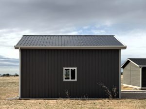 A small, dark gray metal building with a gable roof and a single window on one side, standing on dry, grassy ground under a cloudy sky.