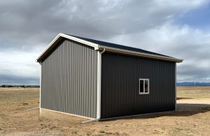 A small, gray metal shed with a gable roof and a single window stands alone on a flat dirt field under a cloudy sky.