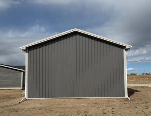 The image shows the side view of a gray metal building with a peaked roof, set on a bare dirt lot under a partly cloudy sky.