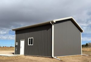 A small gray metal building with a white door, window, and gutter system, standing on a dirt lot under a partly cloudy sky.