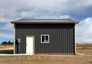 A small, gray metal building with a sloped roof, one white door, and a single window, set on bare ground under a partly cloudy sky.