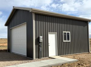 A metal garage with a large white roll-up door, standard door, and window stands on a gravel lot under a partly cloudy sky.