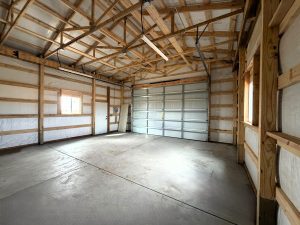 Interior of an empty garage with exposed wooden beams, concrete floor, a large metal overhead door, and two windows letting in natural light.