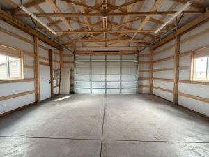 Empty garage interior with exposed wooden beams, insulated walls, concrete floor, two windows, a side door, and a large closed overhead garage door.