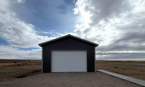 A dark gray metal garage with a closed white door stands on a gravel lot under a partly cloudy sky in a flat, open landscape.