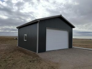 A dark gray metal garage with a white roll-up door and a single window, situated on a gravel driveway in an open, rural landscape under a cloudy sky.
