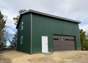 A green metal building with a white door and a brown garage door sits on a sandy lot, surrounded by trees under a cloudy sky.