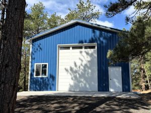 Blue metal building with a large white overhead garage door, a white entry door, and a window, surrounded by pine trees under a partly cloudy sky.