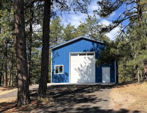 A blue metal building with a large white garage door and a small window, surrounded by tall pine trees on a sunny day.