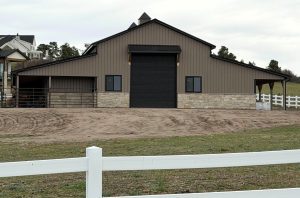 A large barn with a stone lower facade, metal siding, black garage door, and fenced areas sits on a dirt driveway with a white rail fence in front.