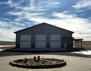 A large metal building with three garage doors stands behind a circular garden bed with potted plants under a partly cloudy sky.