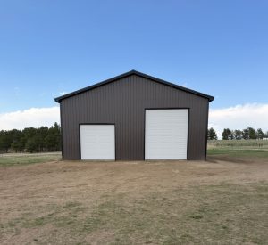 Large brown metal building with two white roll-up garage doors, situated on a dirt lot with trees and blue sky in the background.