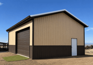 A tan and brown metal storage building with a large garage door and a white entry door sits on a dirt lot under a clear blue sky.