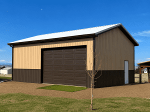 A beige and brown metal building with a large garage door, a side entry door, and a small tree in front, set against a clear blue sky.