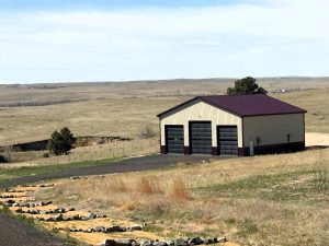 A large metal garage with three bay doors stands on a paved lot amid grassy, open plains under a clear sky.