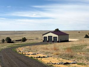 A large metal garage with three bay doors stands on an open grassy plain, next to a curved gravel driveway under a partly cloudy sky.