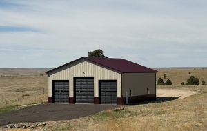 A large metal building with three garage doors stands on a paved area in an open, grassy landscape under a clear sky.