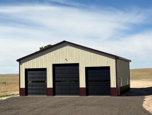 A beige metal garage with three large black doors and a dark red trim, situated on a paved driveway in an open, grassy area under a blue sky.