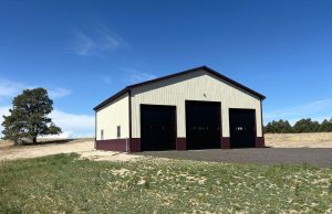 A beige metal building with three large black garage doors stands on a paved area in a grassy, open landscape under a clear blue sky.