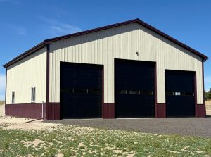 A large metal building with three tall black garage doors and beige and maroon siding, set on a gravel lot with sparse grass.