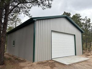 A metal storage building with gray siding, a green roof trim, a white garage door, and a small window on the side, situated among pine trees.