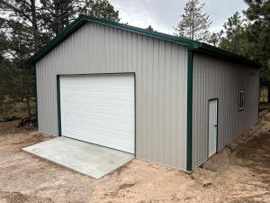 A metal garage with a white roll-up door and a small side entrance, surrounded by trees on bare ground.