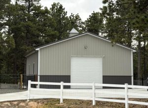 A metal building with a large white garage door stands behind a white fence, surrounded by trees and overcast skies.