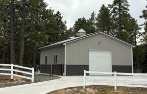 A gray metal building with a white garage door stands beside a paved driveway, surrounded by pine trees and white fencing.