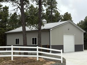 A gray metal building with a white garage door and windows is surrounded by pine trees and a white fence on a concrete driveway.