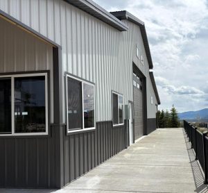 A large metal building with several windows and a garage door sits beside a wide concrete walkway under a partly cloudy sky.