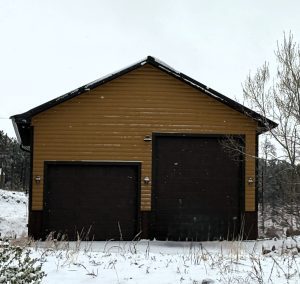 A yellow and brown garage with two large dark doors stands in a snowy, grassy area with bare trees and falling snow.