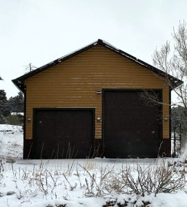 A brown and yellow garage with two large doors stands in a snowy landscape, surrounded by leafless trees and bushes.